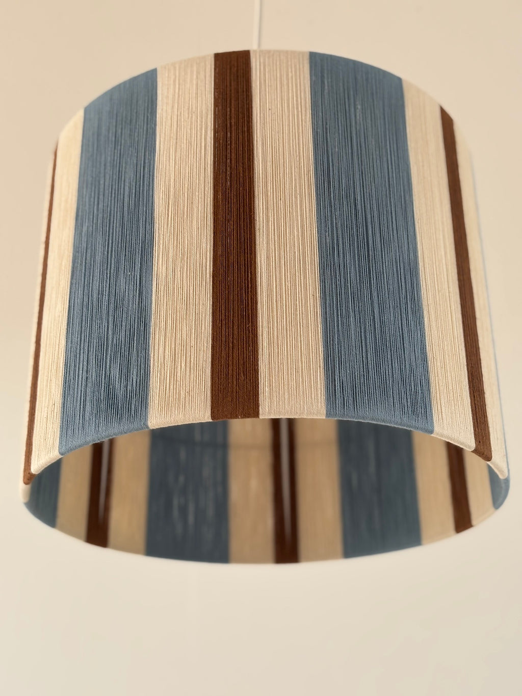 Close-up bottom view of the Cléo Drum pendant light, showing handwoven vertical stripes in cloud blue, natural and fawn yarn