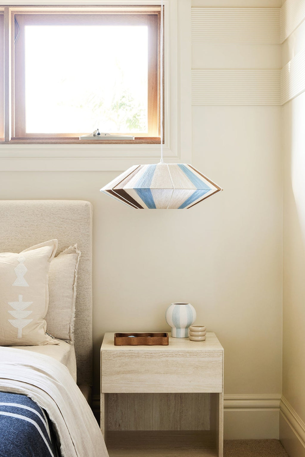 A modern woven pendant light with a blue, white, and brown geometric diamond pattern hangs above a light wood bedside table in a bright, neutral-toned bedroom.